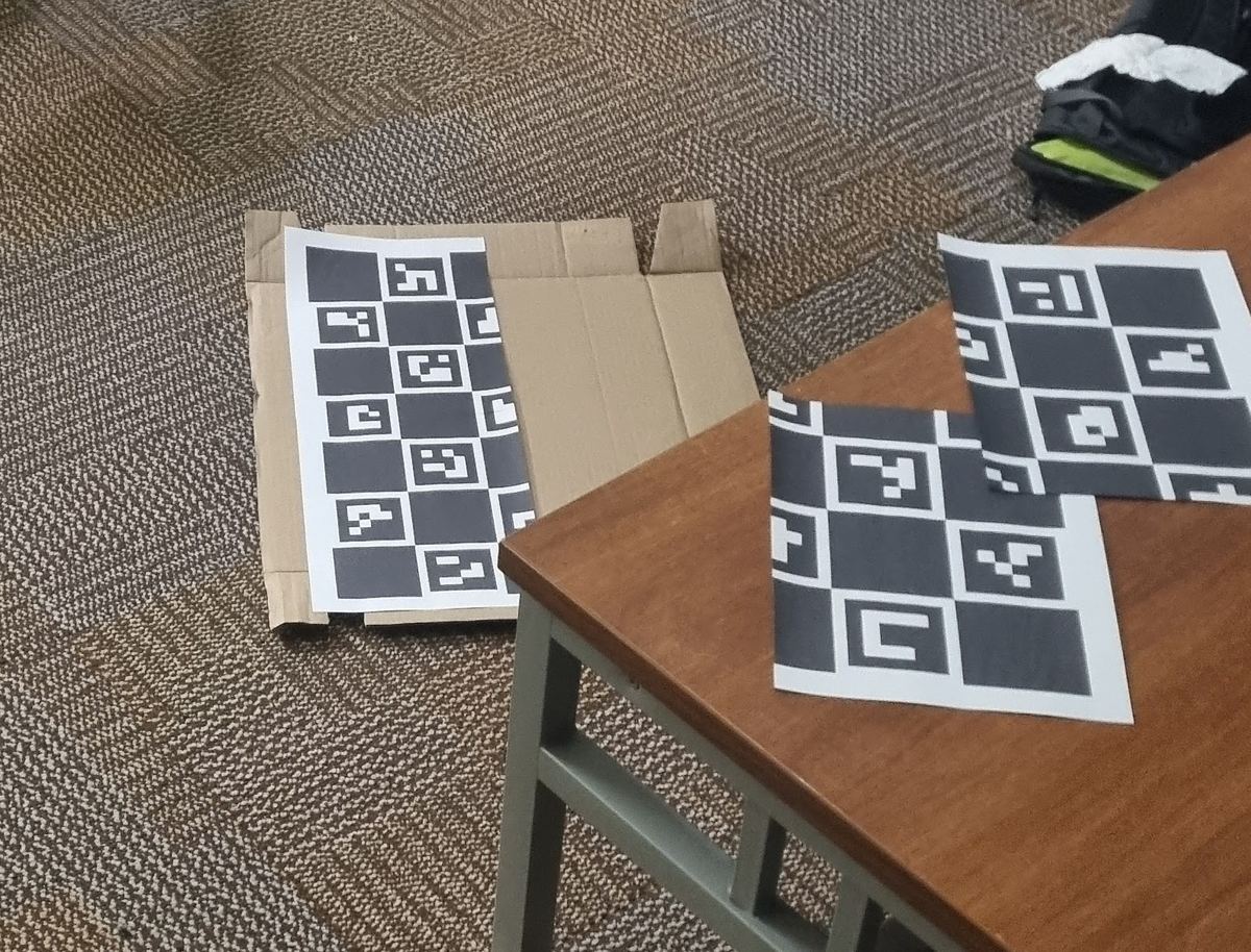 Three printed sheets with a black-and-white checkerboard pattern and small QR-like squares inside it. One sheet lies on a piece of cardboard on a carpet, the other two lie on the corner of a wooden table.