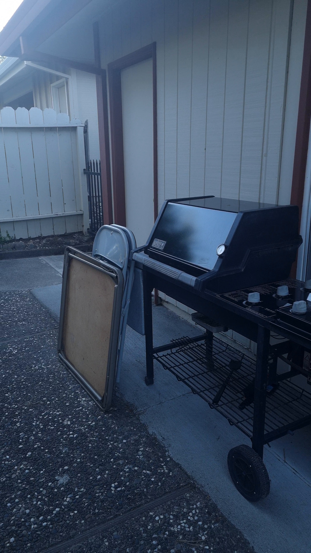 A barbecue and a framed board against a house wall, on a gravel patio.
